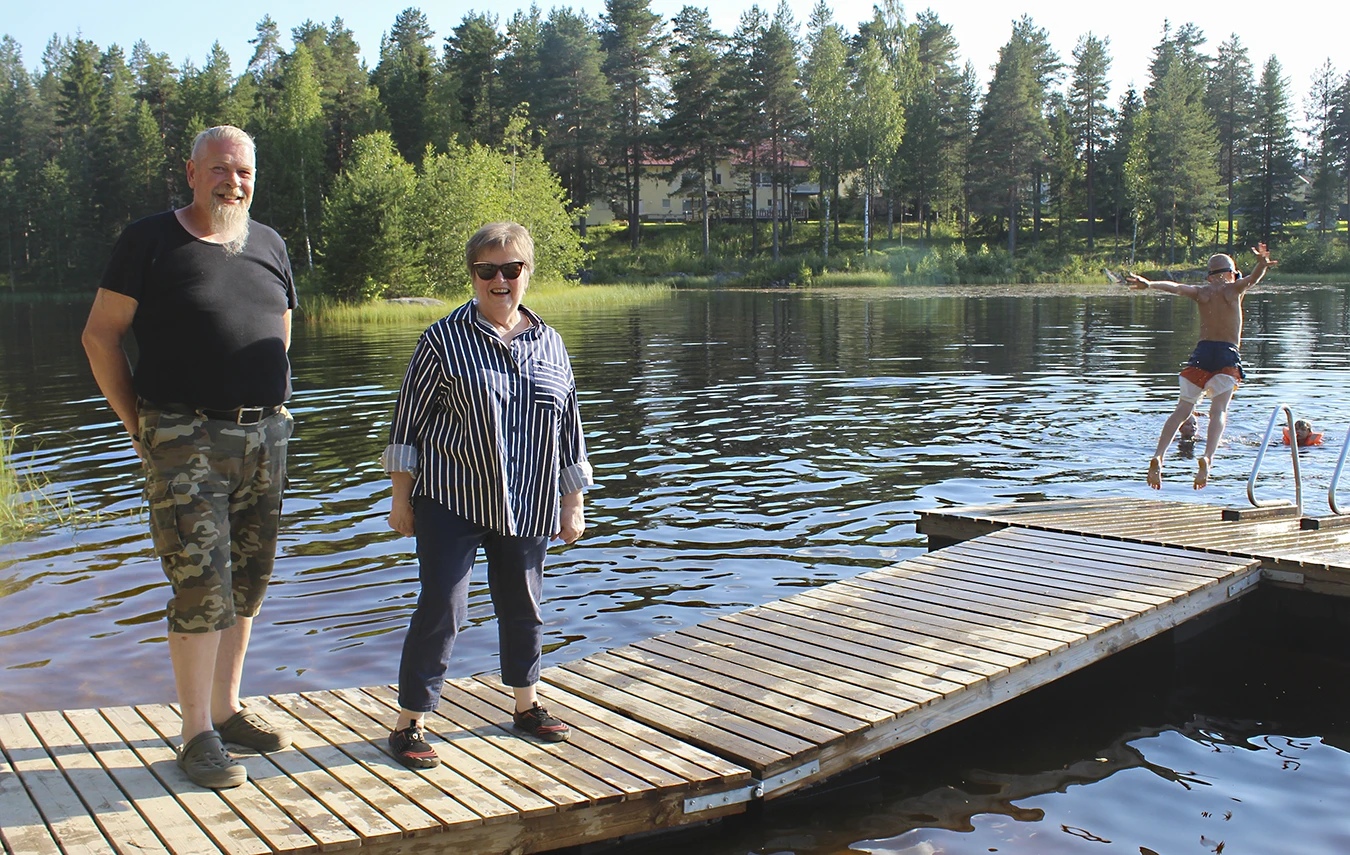 Kaksi ihmistä seisoo laiturilla. Poika hyppää veteen laiturin päästä.